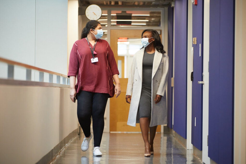 Maryam Ahmad and Kizzmeika Corbett walk down an academic hallway in conversation. They are wearing face masks.