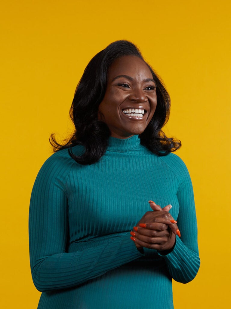 Kizzmekia Corbett wearing a longsleeve, teal turtleneck dress, smiles and looks off camera with her hands clasped.