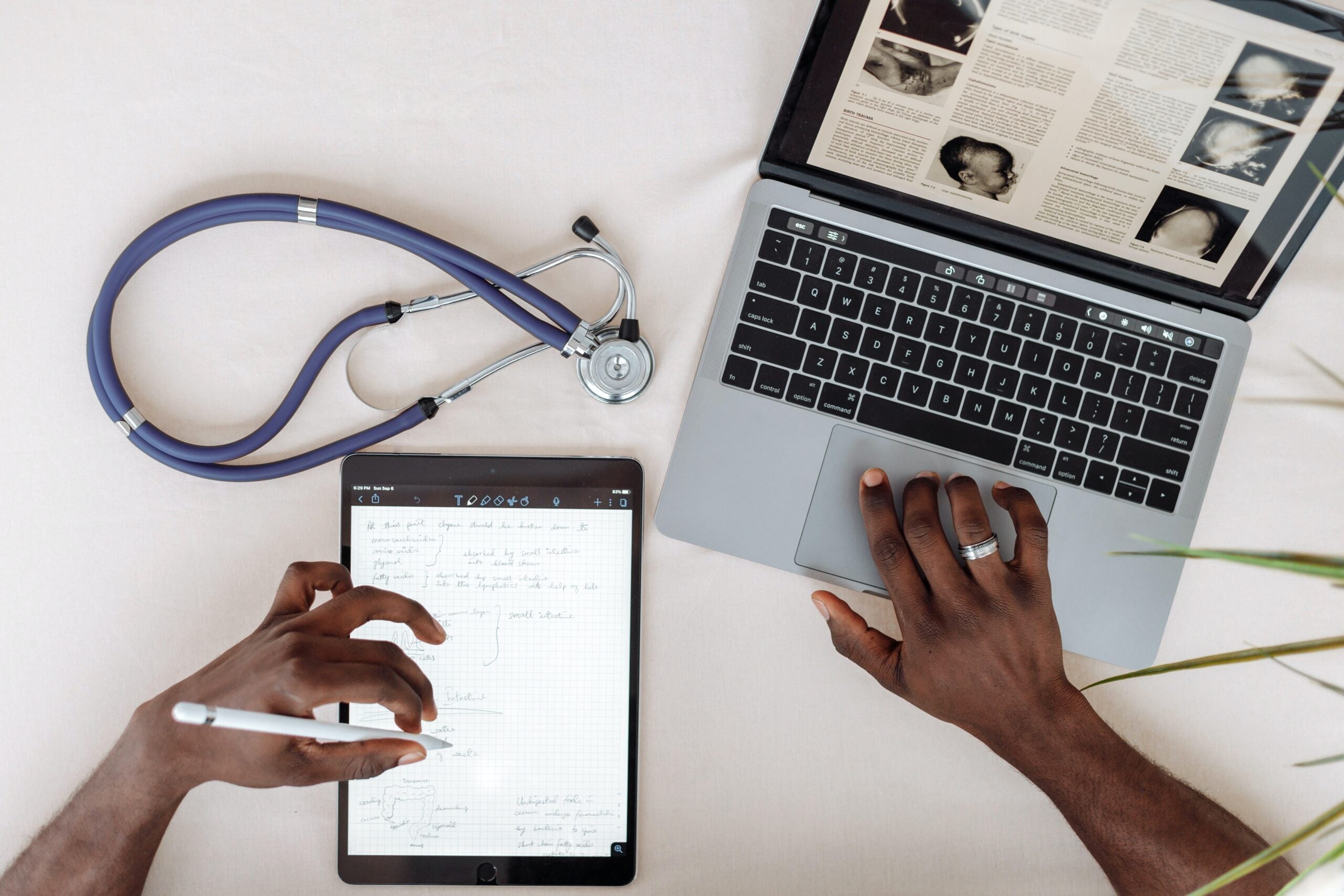 Image of a desk containing a laptop showing a medical article, as well as a stethoscope and a tablet with notes