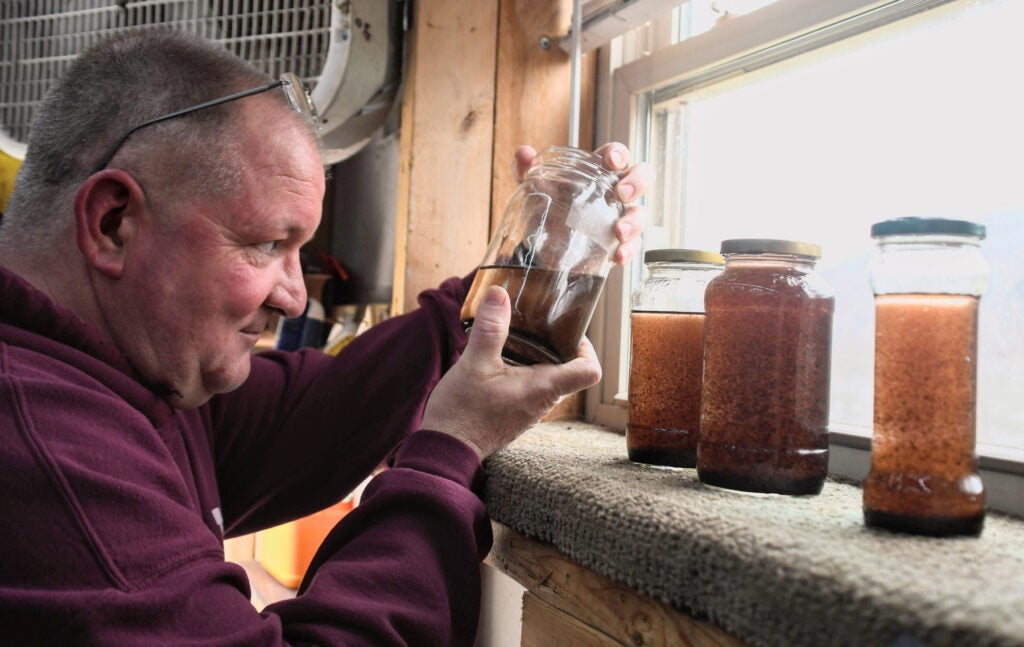 A white man wearing a maroon sweatshirt with glasses on his head inspects a jar of dirty water from a window sill in his basement workroom. Three other jars of dirty water on the sill.