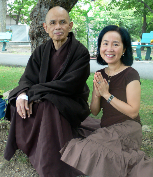 Thich Nhat Hanh and Lilian Cheung squat in a grassy area. Lilian holds her palms together, in a prayerful pose. 