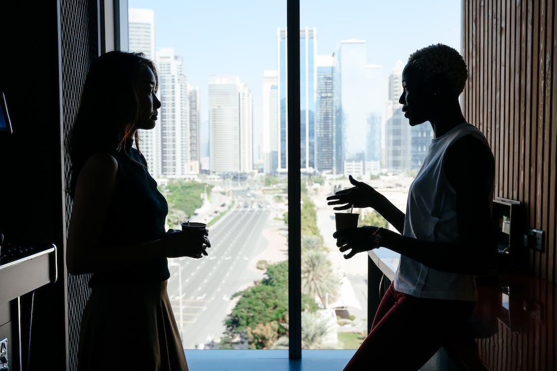 Two women having a serious discussion, holding coffee, and standing in front of a large window in an office.