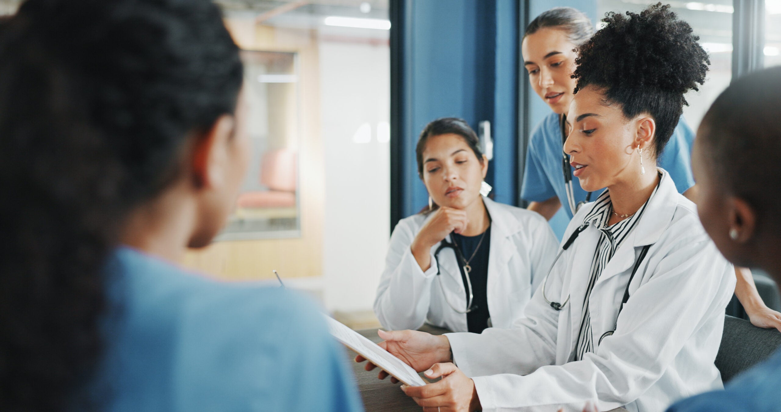 A group of health professionals surround a doctor holding a clipboard seated at a table.