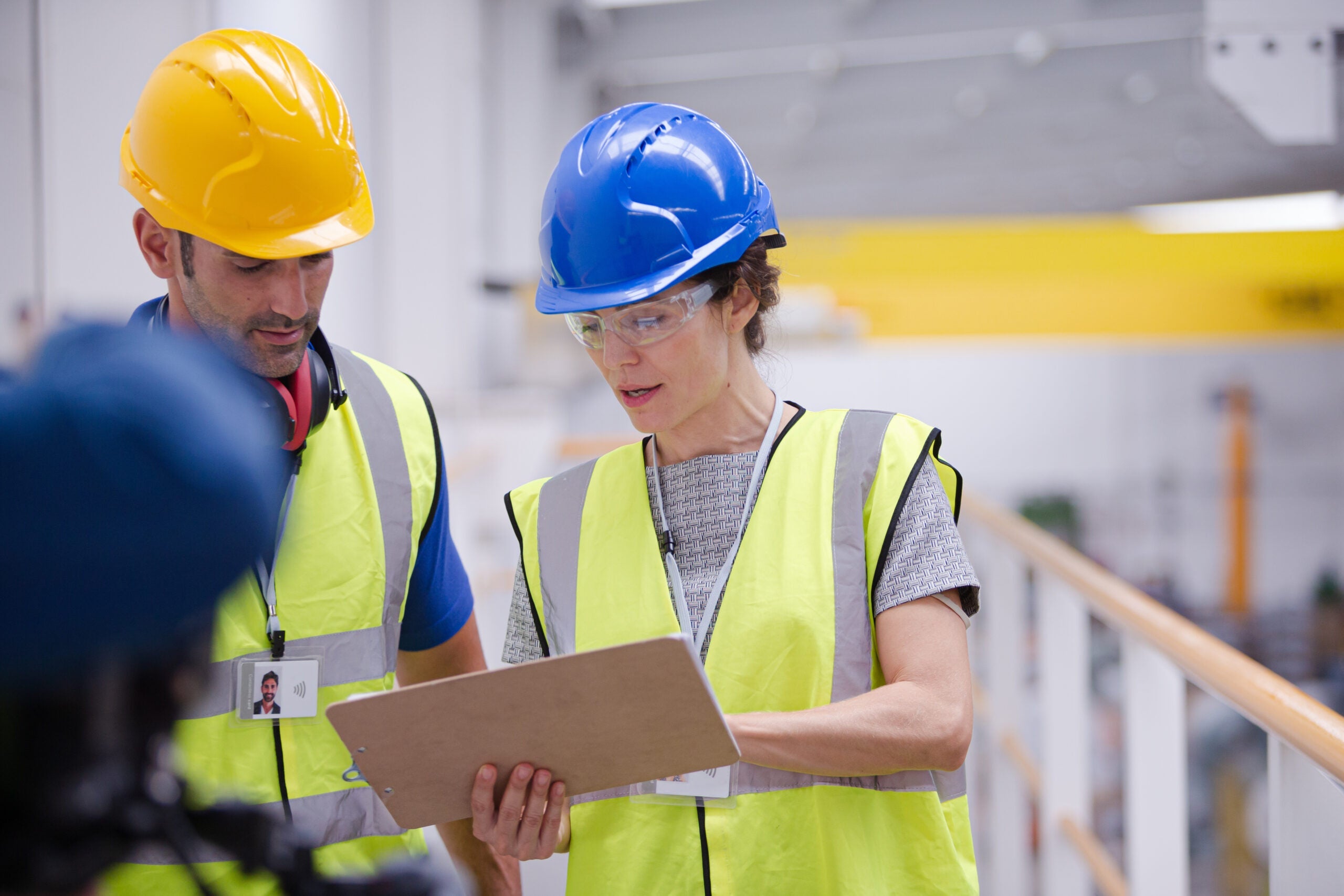 Two people wearing construction vests and hard hats discussing information on a clipboard, inside of a factory