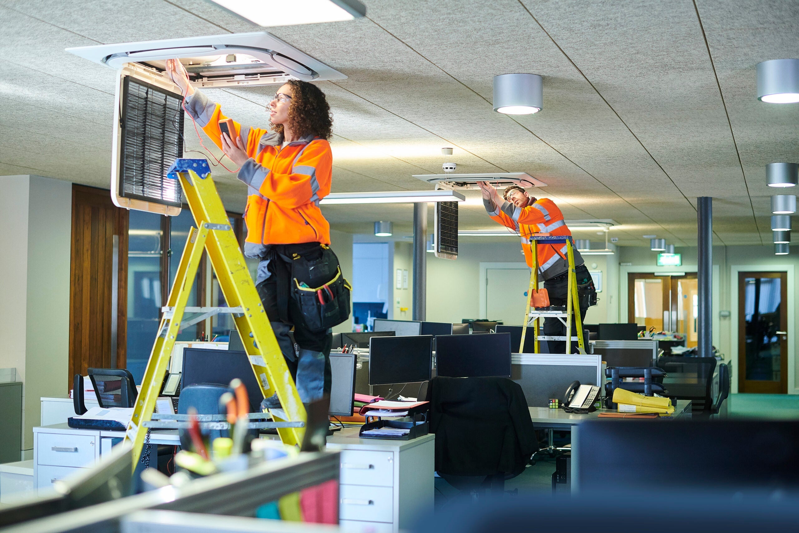 Two professionals wearing safety gear standing on ladders as they examine the air vents in an office building