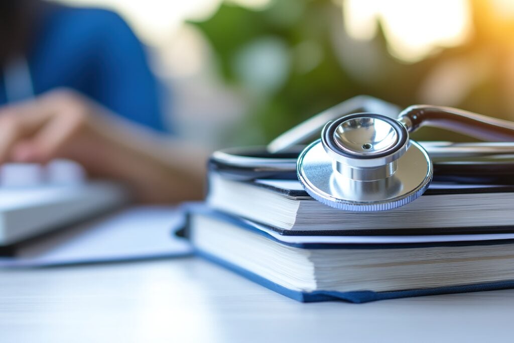 A close-up of a stethoscope resting on top of a stack of open books, with a blurred figure typing on a keyboard in the background. The scene is brightly lit, suggesting a professional or educational setting.