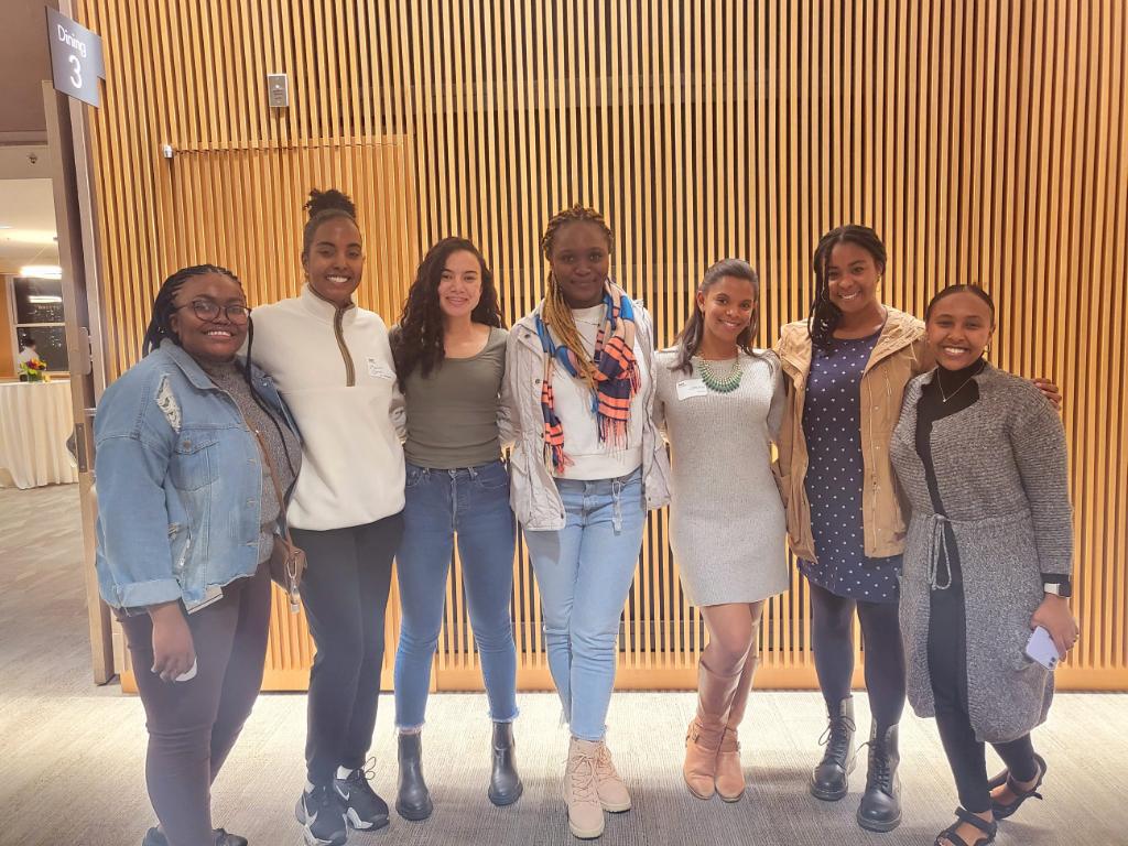 Seven students standing in front of a tan wall.