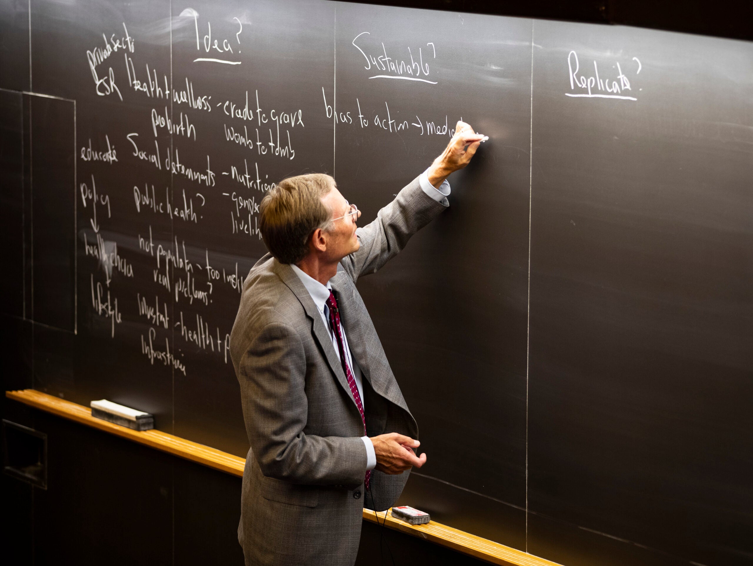 A faculty member in a suit and tie writes on a blackboard in a classroom