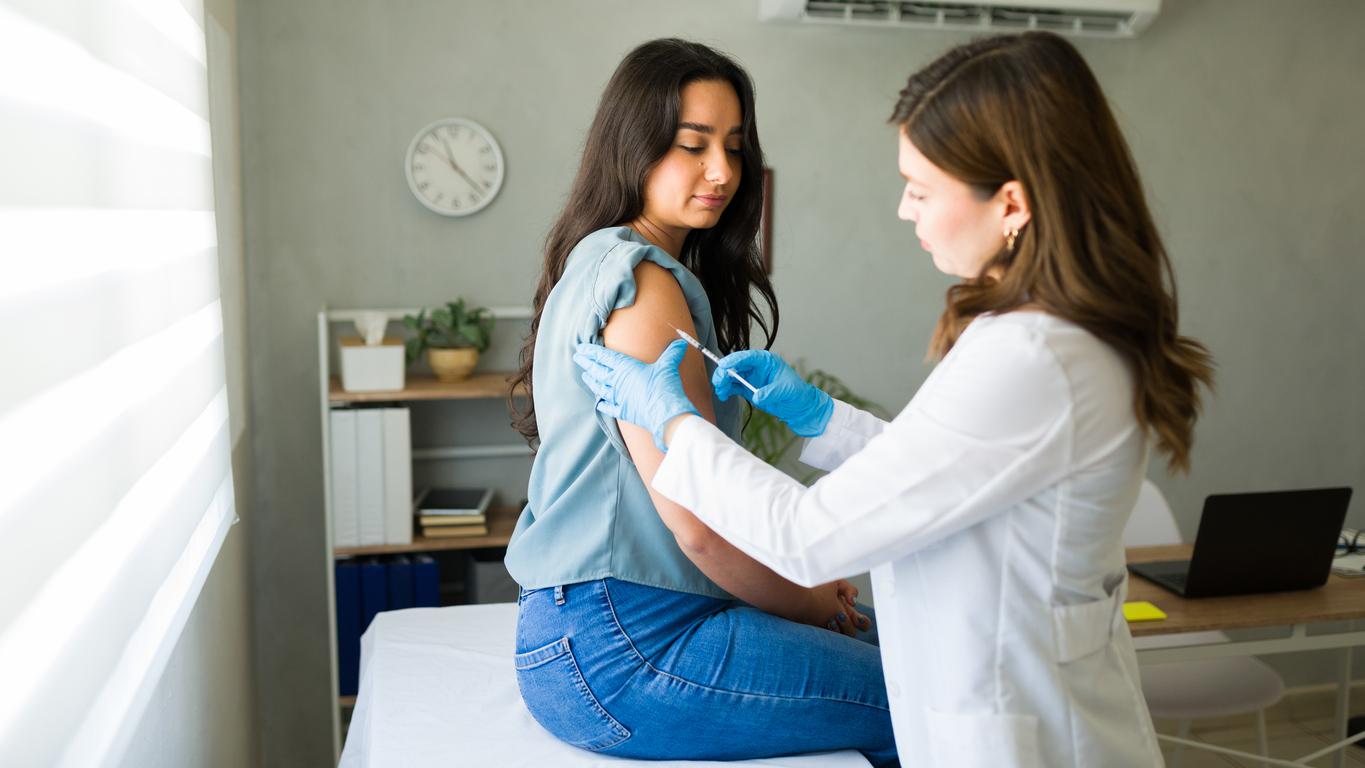 A patient sits on a hospital exam room cot while a nurse administers a vaccine in her arm.