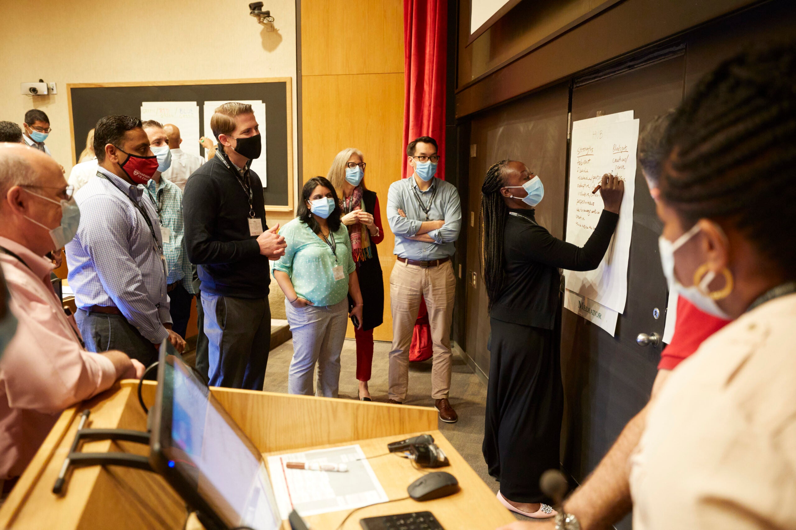 A group of students standing near the front of a Harvard Chan classroom, wearing masks, and watching a student write on a whiteboard