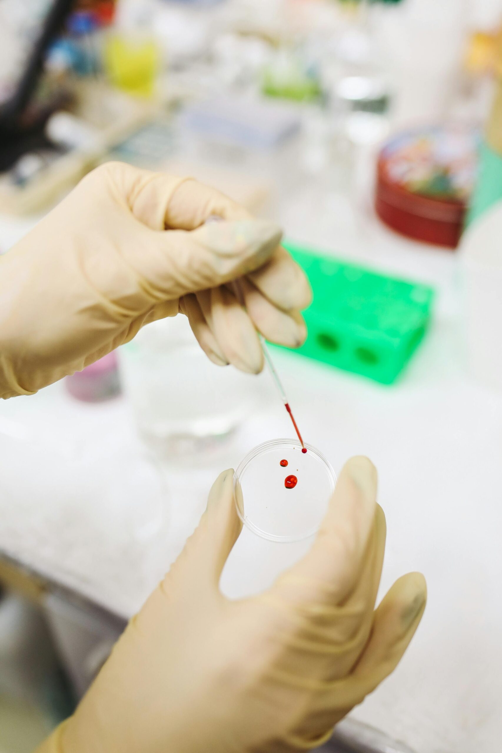 A gloved hand holds a petri dish and pipette containing a red liquid.