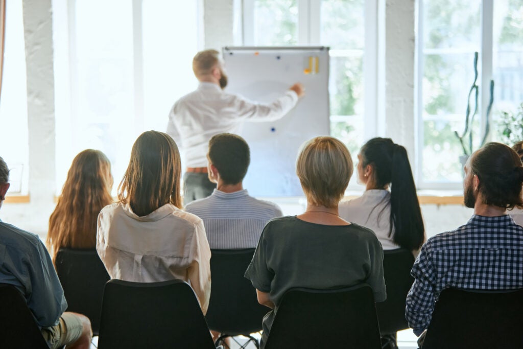 Group of people seated in a classroom watching a man give a presentation at a whiteboard in a bright, sunlit room.