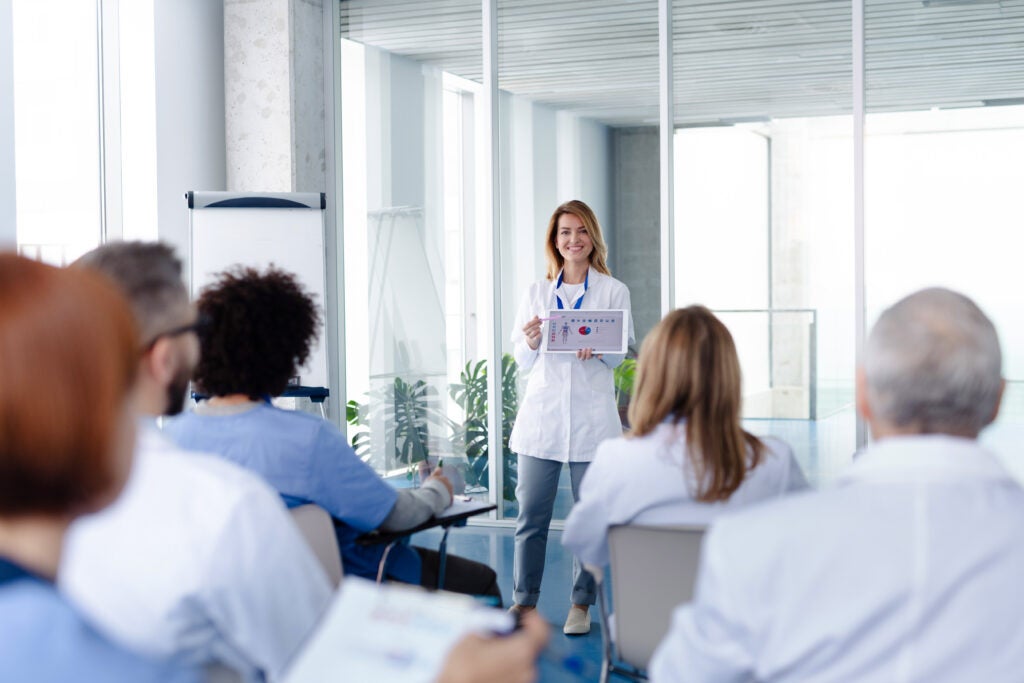 
A female medical professional in a white coat stands in front of a group, holding a chart with graphs. She appears to be giving a presentation in a bright, modern conference room with large windows. Her audience consists of other healthcare workers, seated and taking notes.