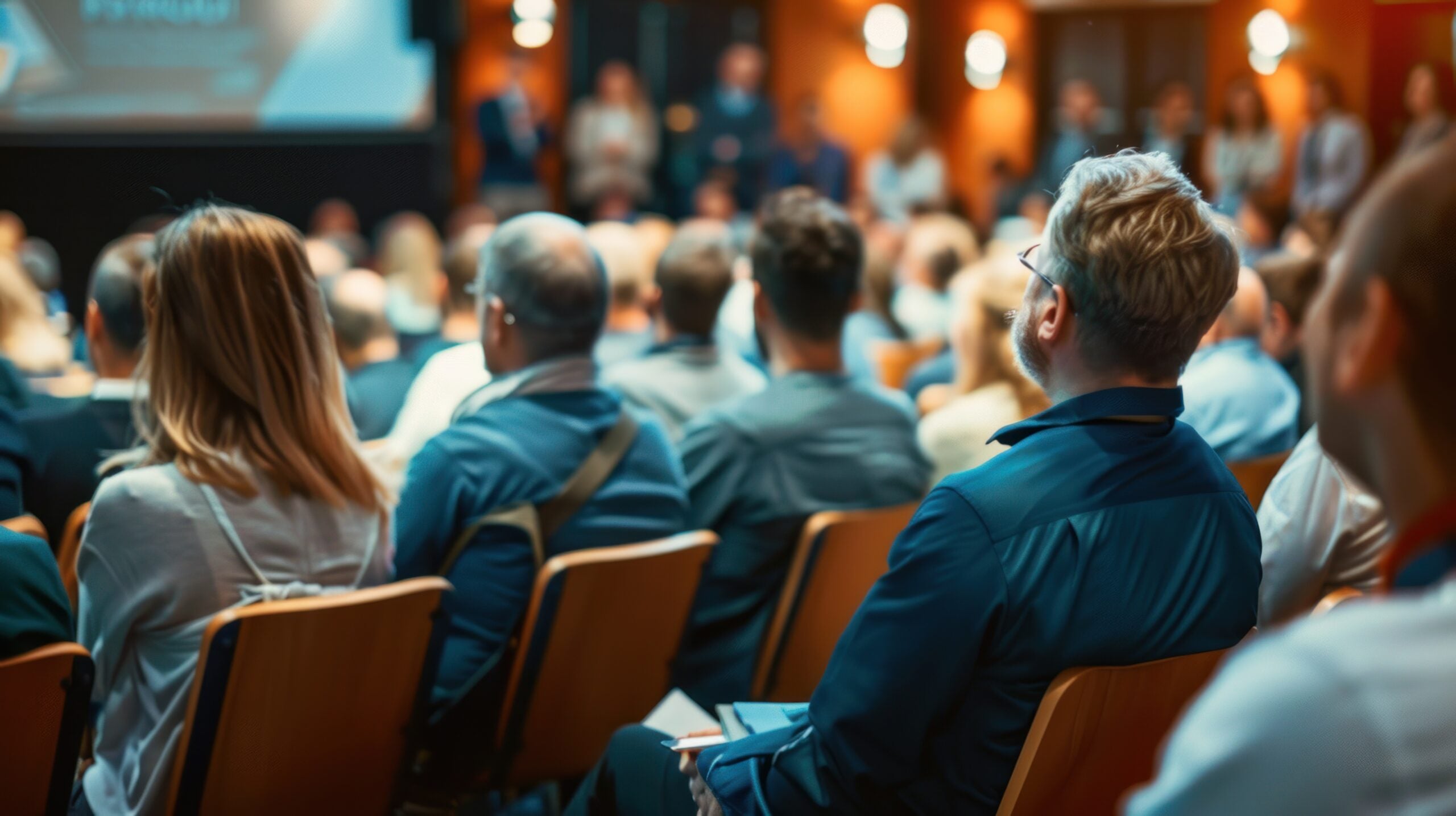 A large group of people seated in rows of wooden chairs, attentively watching a presentation or lecture in a well-lit auditorium or conference room. The focus is on the audience members from behind, while a blurred projection screen and several standing figures are visible in the background.