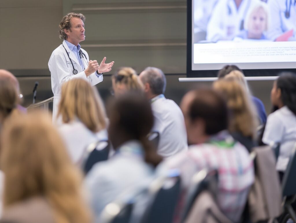 Male doctor in a white coat with a stethoscope speaks to an audience of healthcare professionals at a conference, with a presentation slide in the background.