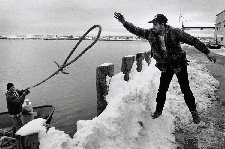 Black and white photo of a man standing on a snow-covered dock reaching out to catch a rope thrown by another man on a boat in a harbor.