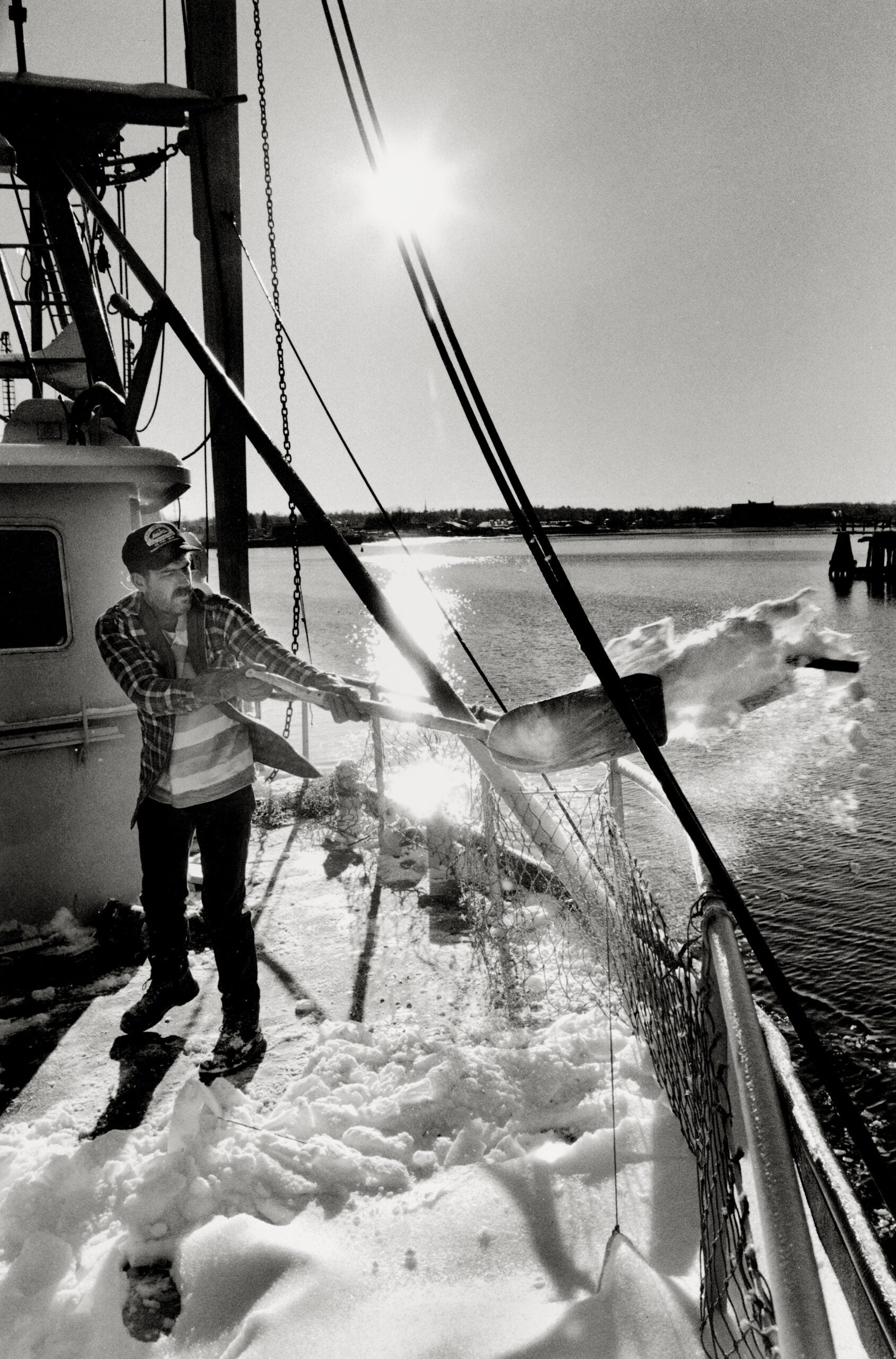 Black and white photo of a man on a boat using a shovel to clear snow, with the sun shining brightly in the background over the water.