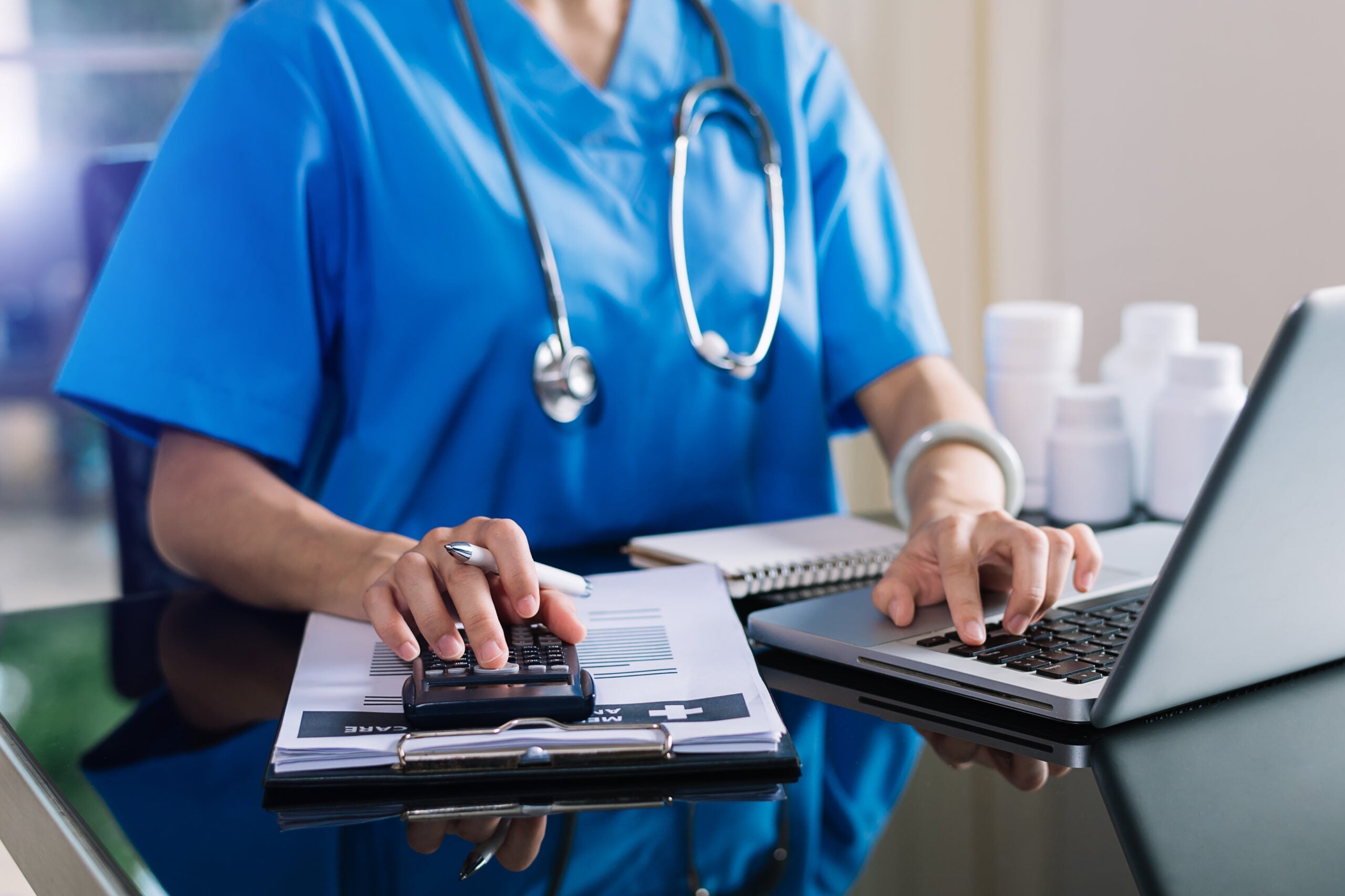 Close-up image of a doctor simultaneously typing on a laptop and entering numbers into a calculator