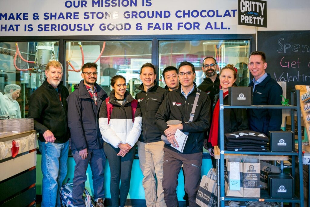 Group of people standing together inside a chocolate factory, posing in front of a sign that reads 'OUR MISSION IS TO MAKE & SHARE STONE GROUND CHOCOLATE THAT IS SERIOUSLY GOOD & FAIR FOR ALL.'