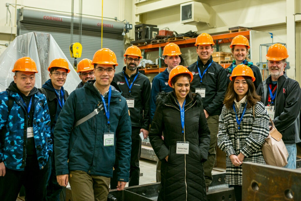 Group of people wearing orange hard hats and lanyards standing together in an industrial or construction setting.