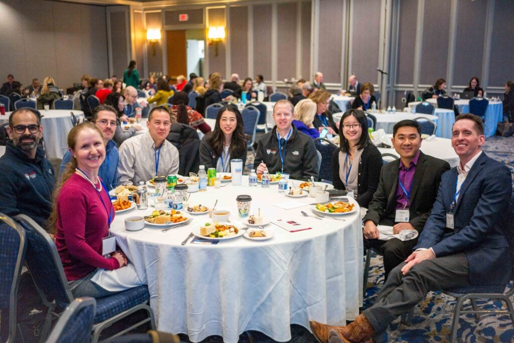 Group of people sitting at a round table with food, attending a conference or event in a large dining hall.