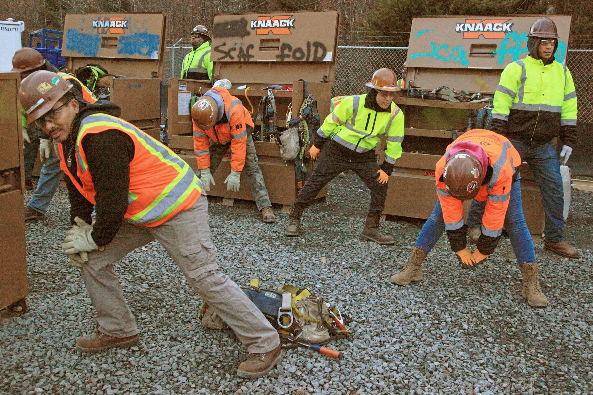 Construction workers wearing high-visibility jackets and hard hats are performing stretches or warm-up exercises on a gravel surface in front of open metal storage boxes labeled "KNAACK." Various tools and equipment are visible inside the boxes.