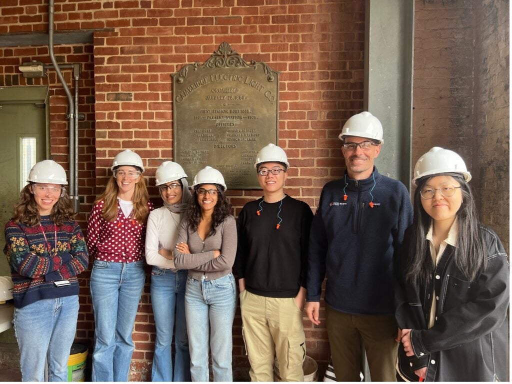 A group of people wearing hard hats and safety glasses standing in front of a historical plaque on a brick wall. They are smiling and appear to be on a tour or site visit at an industrial or historical facility.