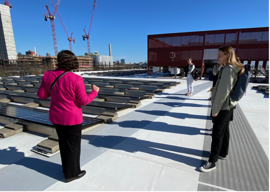 A group of people stand on a rooftop with solar panels, listening to a woman in a pink jacket who is addressing them. The backdrop includes construction sites with cranes, modern buildings, and a clear blue sky, indicating the discussion is likely related to renewable energy or sustainable infrastructure in an urban setting.