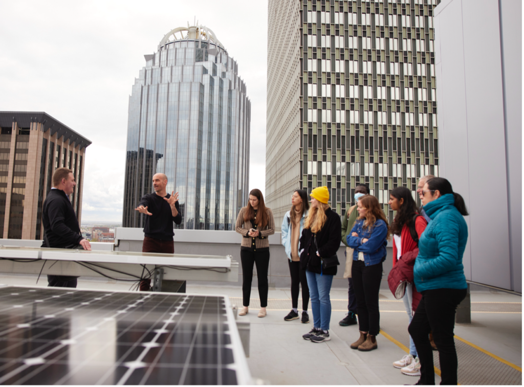 A group of people standing on a rooftop with solar panels, listening to two individuals speaking. The background includes tall, modern buildings, indicating an urban environment. The setting suggests a discussion or tour related to renewable energy or sustainable practices.