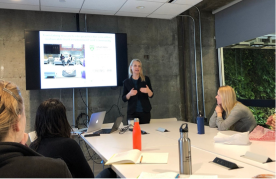 A woman stands at the front of a conference room, giving a presentation to a group of seated individuals. A screen behind her displays a slide with text and images. The setting is professional, with notebooks, laptops, and water bottles on the table, indicating an engaged audience in a meeting or workshop.