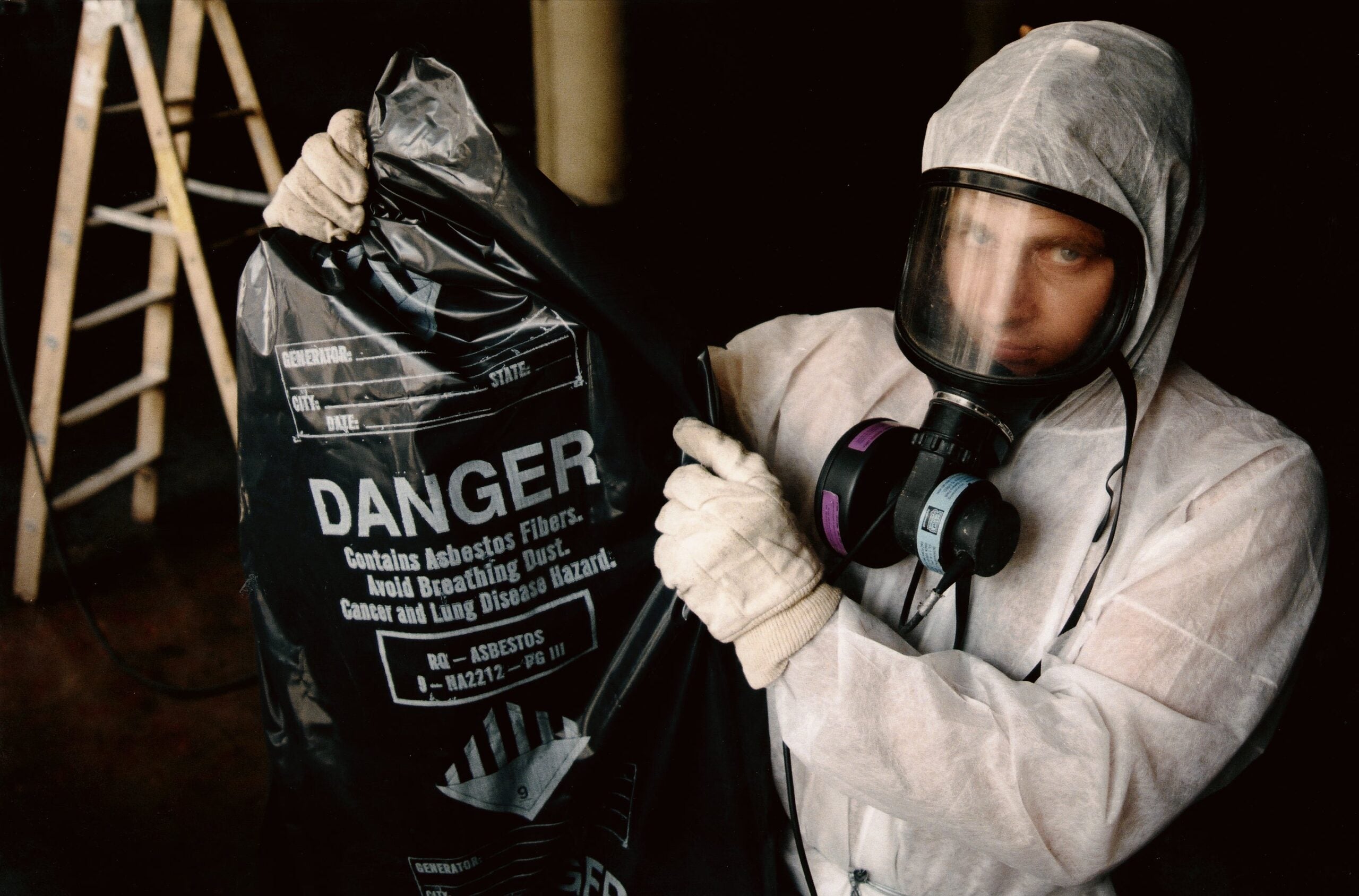 A person wearing a white protective suit, gloves, and a respirator mask holds a black plastic bag labeled with a "DANGER" warning about asbestos. A ladder is visible in the background. The bag's text warns about asbestos fibers, instructing to avoid breathing dust due to cancer and lung disease hazards.