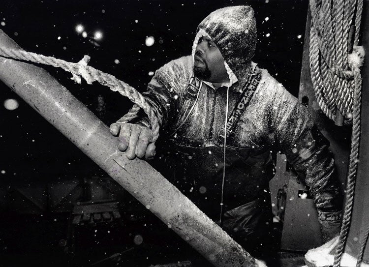 Black and white image of a person dressed in heavy protective gear, covered in snow or frost, likely engaged in work on a ship. They are grasping a large pole or beam, with ropes visible around them. The scene appears to be in challenging, snowy conditions, highlighting the harshness of the environment.