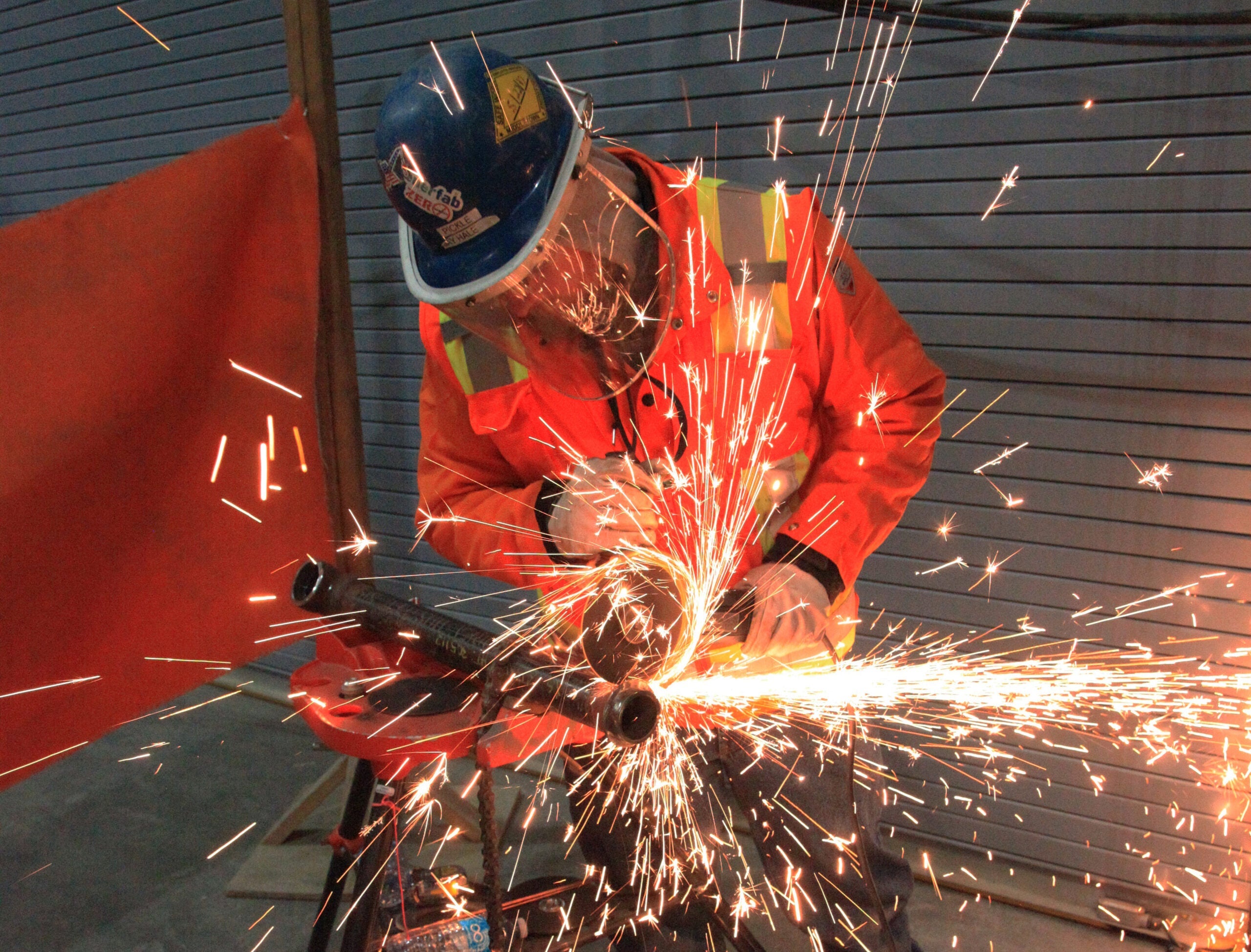 A construction worker wearing a blue hard hat, safety goggles, and an orange high-visibility jacket is using a power grinder on a metal pipe, creating bright sparks that fly in all directions. The worker is in a closed room with red safety barriers and a grey shuttered background.