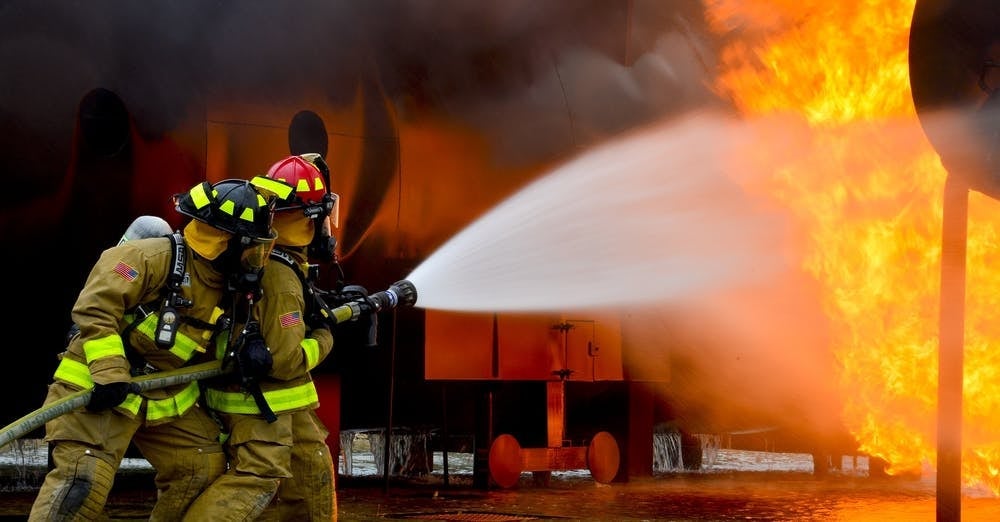 Two firefighters in full gear, including helmets and breathing apparatus, are using a hose to spray water at a large fire. The flames are intense, and thick black smoke is rising in the background. The scene is set in an industrial area with metal structures partially visible.