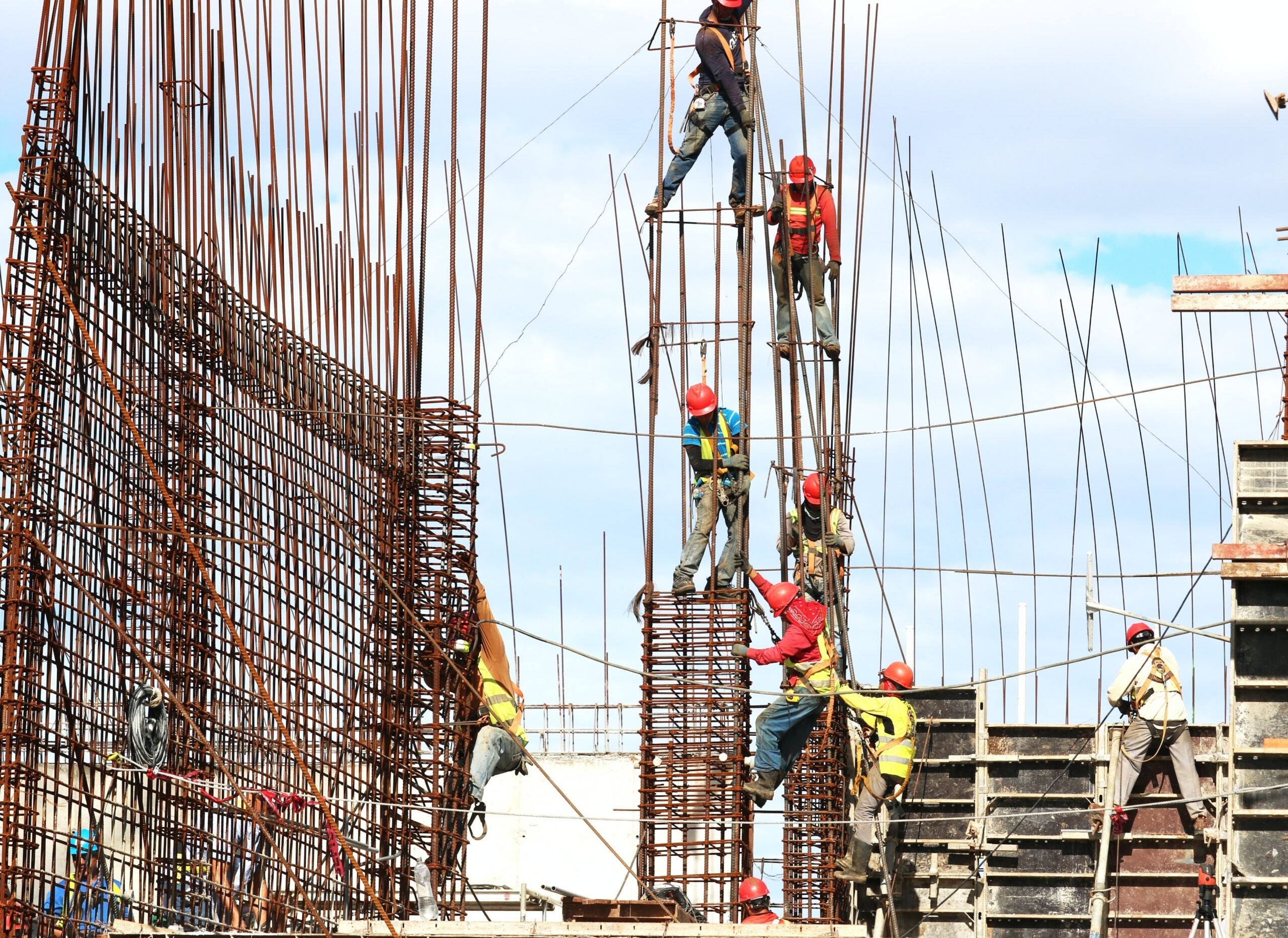 Construction workers wearing safety gear, including red hard hats and high-visibility vests, are working on a building site. They are climbing and securing steel rebar structures, preparing for concrete pouring. The scene is busy with multiple workers at different heights, emphasizing the scale and complexity of the construction project. The sky is clear with a few clouds in the background.