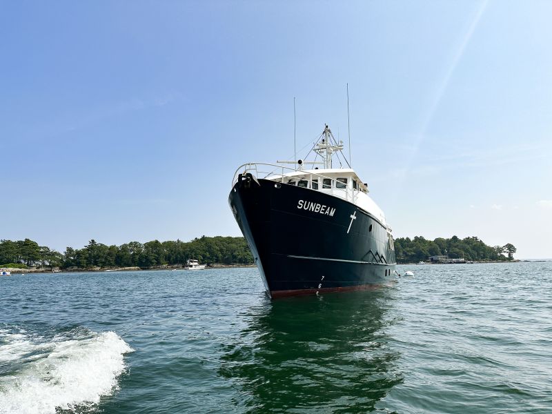 A large black boat named "SUNBEAM" anchored in the water with trees and a shoreline visible in the background under a clear blue sky.