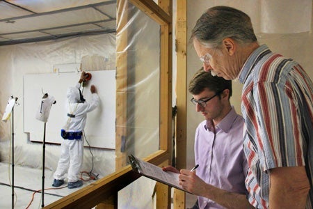 Two individuals are observing and taking notes on a clipboard while a person in full protective gear, including a white coverall suit and a respirator, uses a power tool on a wall inside a controlled environment. The observers are separated from the worker by a transparent plastic barrier.