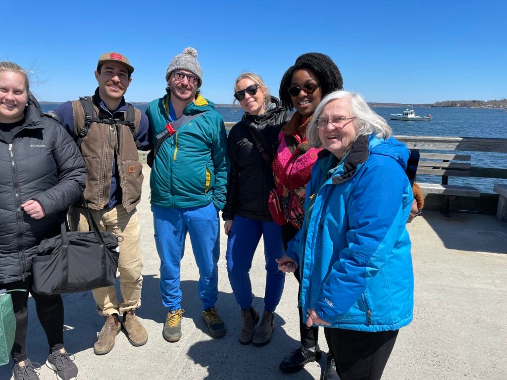 A group of six people standing on a pier by the water, bundled up in winter jackets and smiling towards the camera, with a small boat visible in the sea and a clear blue sky in the background.