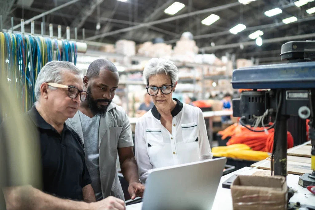 Three people standing together in an industrial workshop, looking at a laptop. The background features an array of tools, equipment, and materials, indicating a collaborative work environment in a manufacturing or engineering setting.