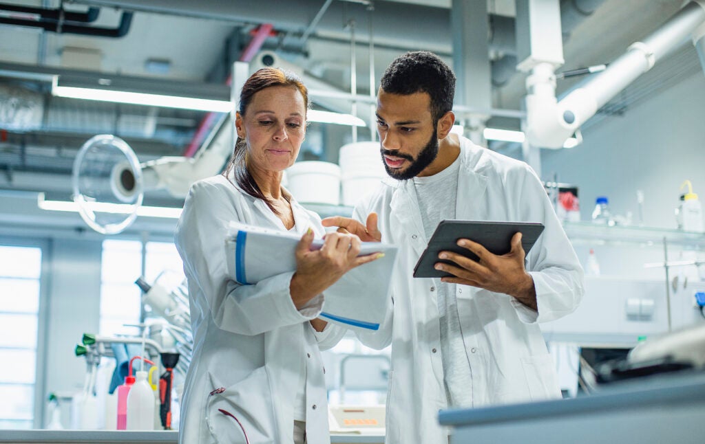 Two scientists in lab coats stand in a modern laboratory discussing their findings. One holds a clipboard, and the other uses a tablet, surrounded by lab equipment and scientific instruments, highlighting a collaborative and high-tech research environment.