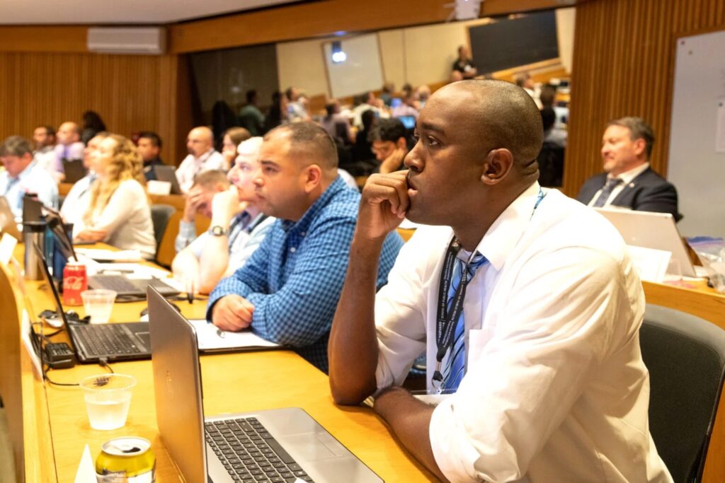 People seated in a conference room, attentively listening to a presentation. They have laptops open in front of them and are taking notes, indicating a focused and engaged learning or professional environment.