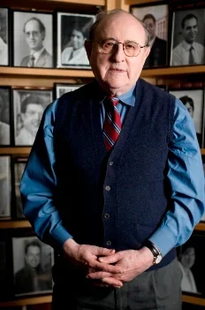 Dr. Bernard Lown, standing with hands folded in front of a wall of photos.