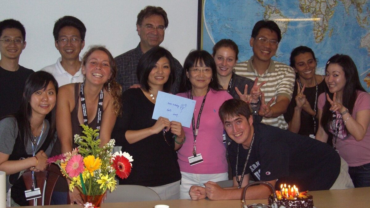 A group of twelve people are gathered around a table, smiling and posing for the camera. A woman in the center holds a card, possibly for a celebration. On the table, there are a bouquet of colorful flowers, a cake with lit candles, and some cups and utensils. A world map is displayed on the wall in the background. The mood is joyful and festive.