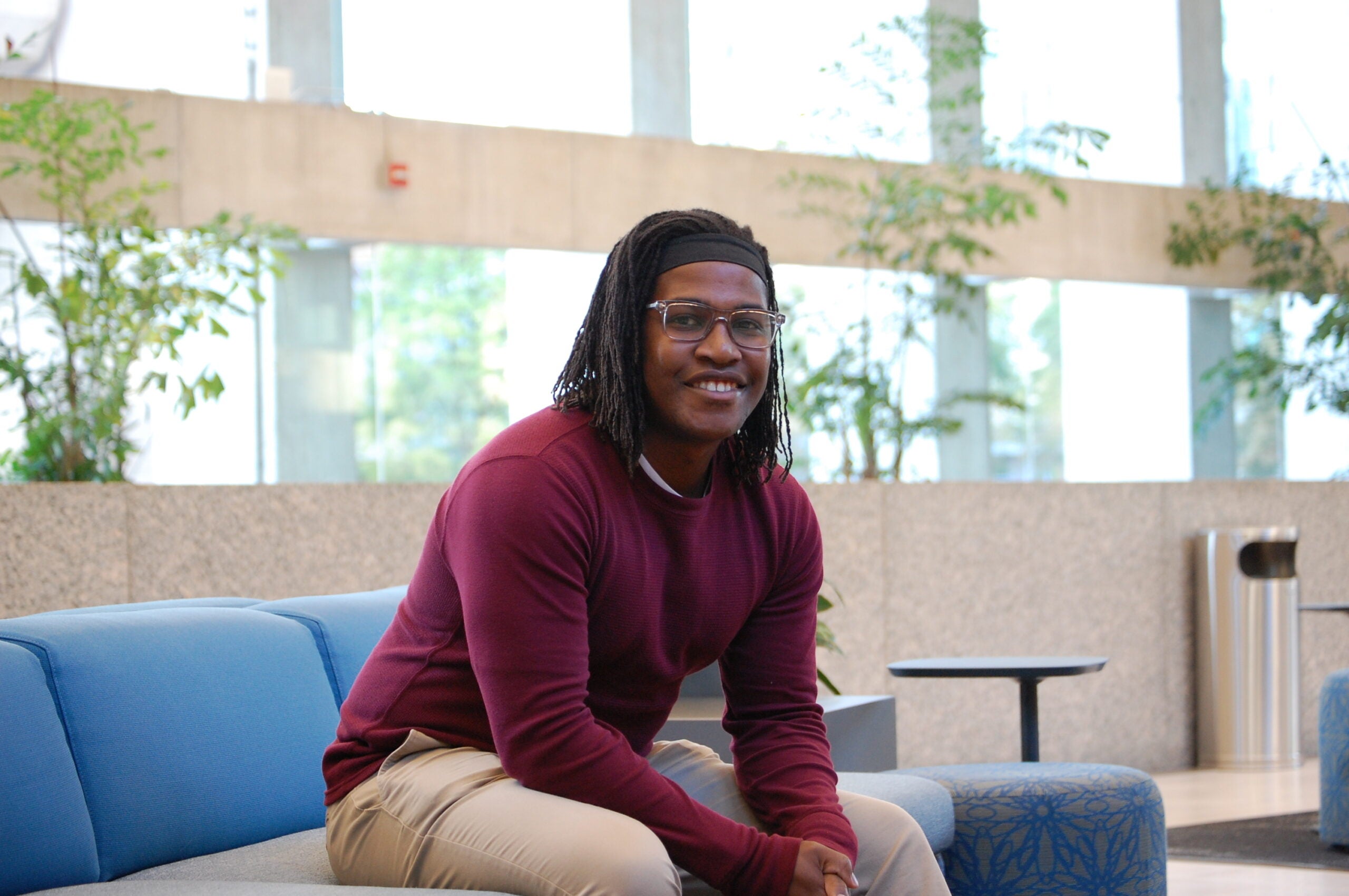 a headshot of Tinashe Michael Tapera. He is wearing a red sweater and sits on a blue and beige couch with windows behind him