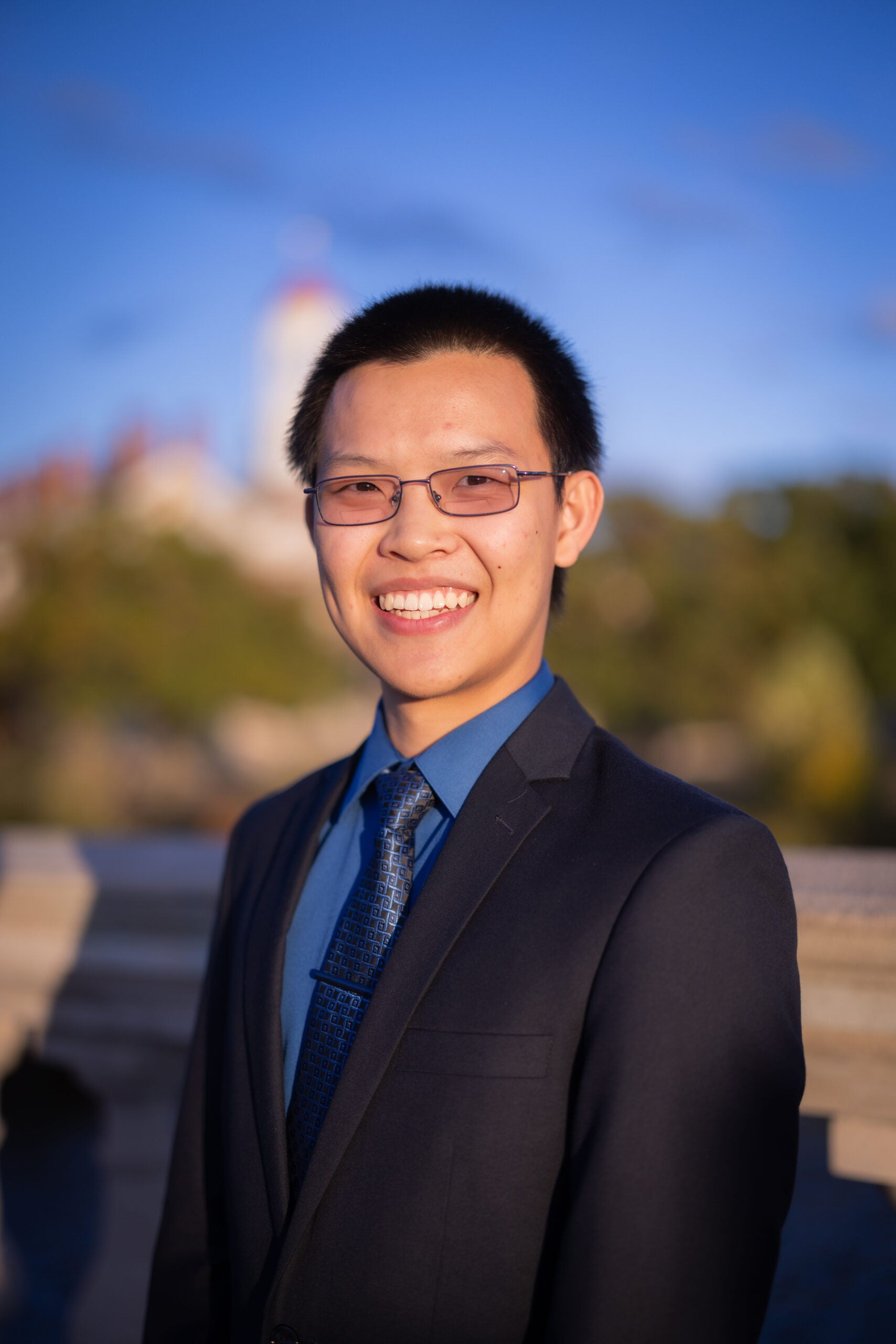 a headshot of Eric Tong. he is outside, and the background is blurred. the sky is blue and there is a lighthouse and hills behind Eric. Eric wears glasses and is smiling at the camera. He wears a dark blue dress shirt with a dark colored tie under a black suit jacket