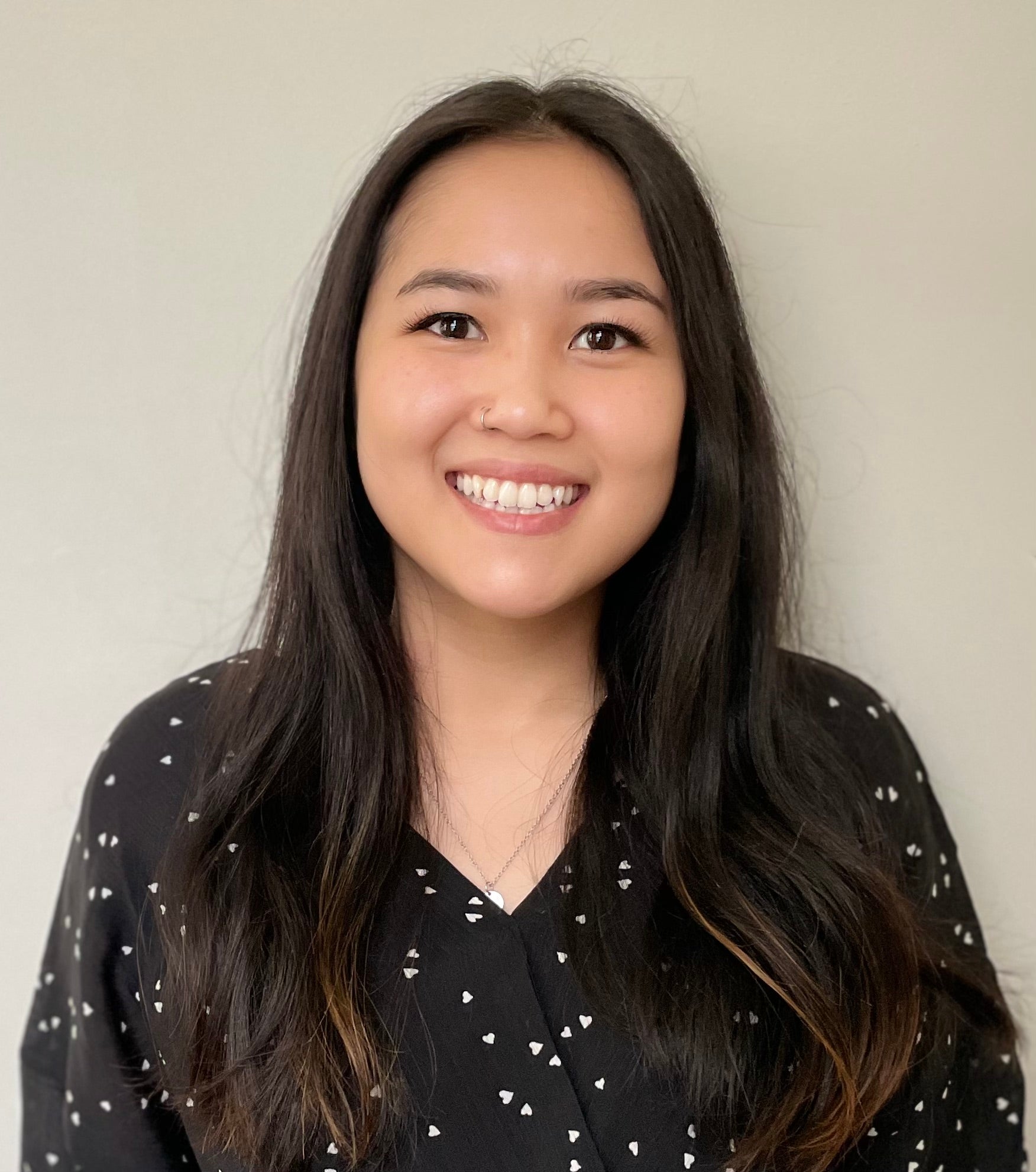 a headshot of Dr. Sophia Le. she is smiling at the camera and wears a black, long sleeve blouse with white polka dots in the shape of hearts. her hair is past her shoulders and worn down on both sides. she has a nose ring on the viewer's left side and is also wearing a necklace, the pendant partially hidden by her blouse collar.