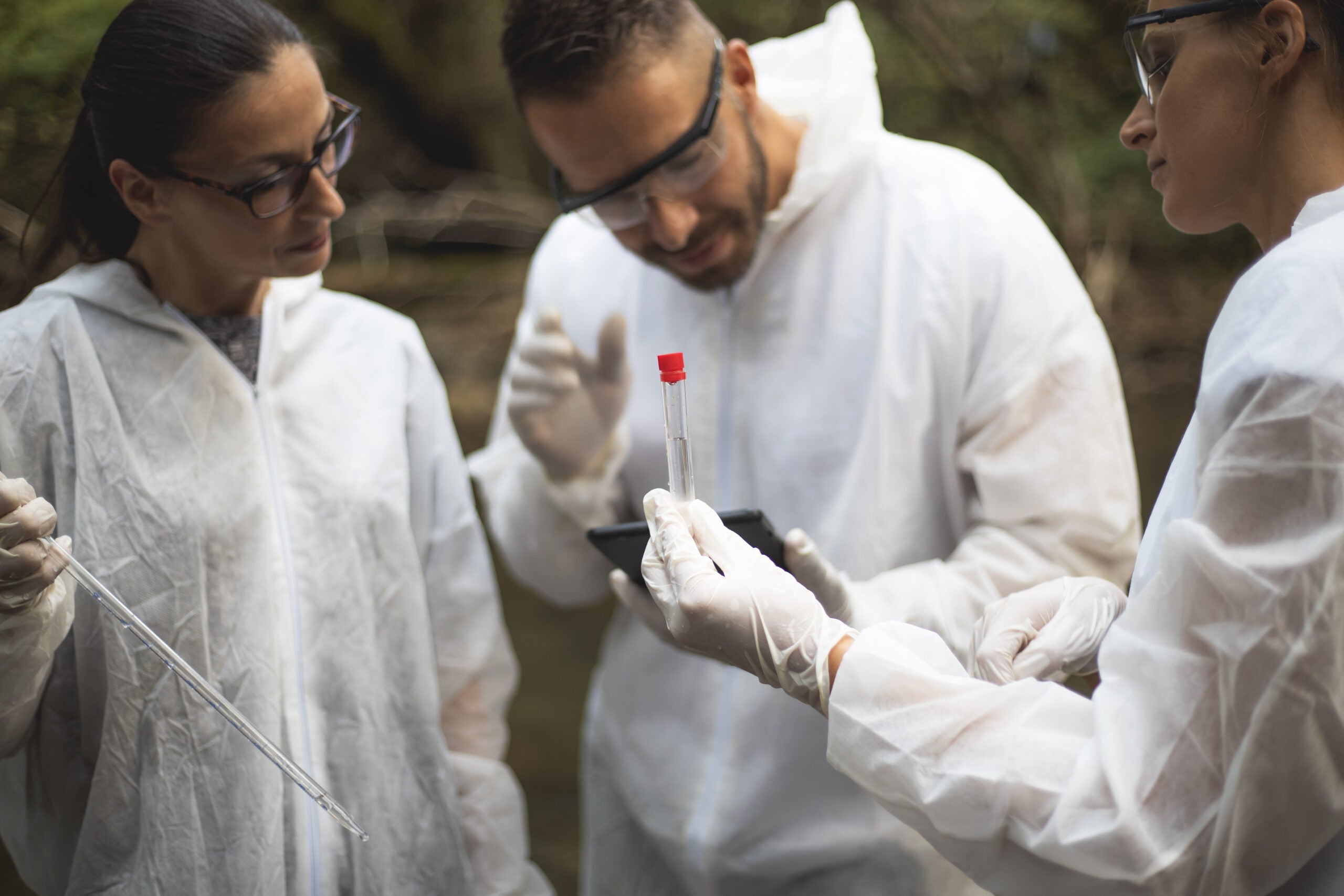 Image of 3 scientists standing outdoors, with one holding a vial of clear liquid