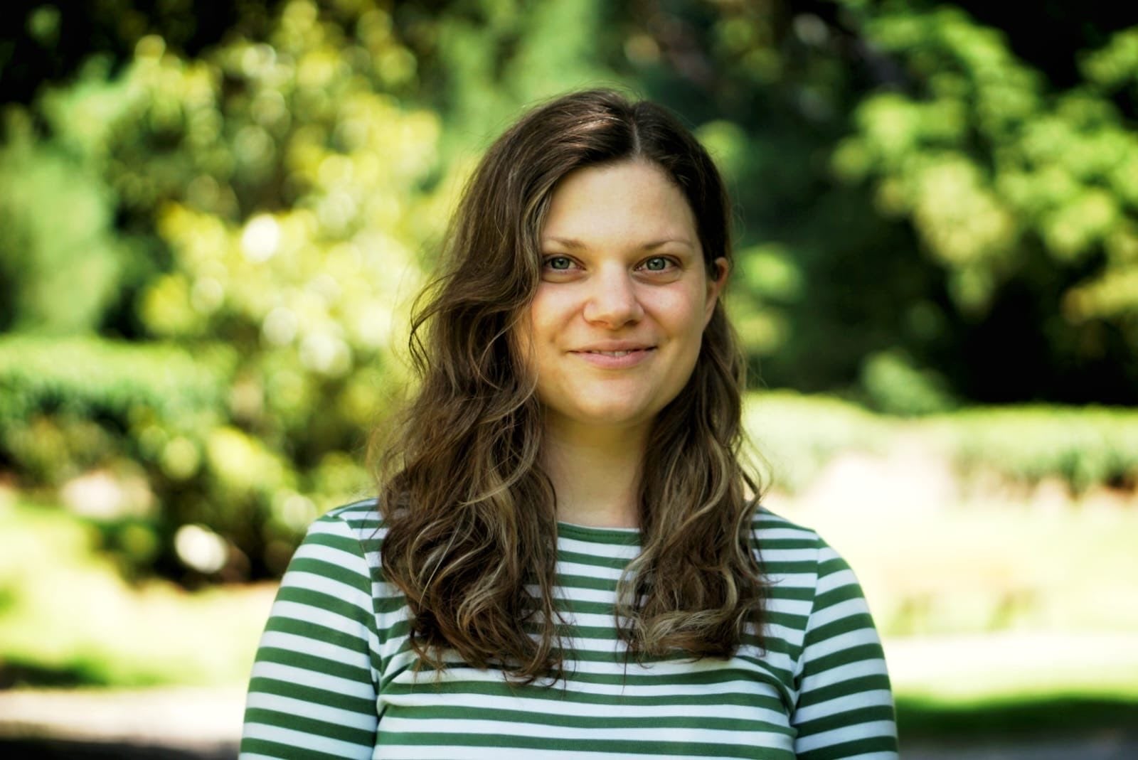 a headshot of Dr. Lisa Thalheimer. She has shoulder-length brown hair, and is wearing a black and white horizontal striped shirt. she smiles at the camera and the background indicates she is outside with green and yellow bushes behind her