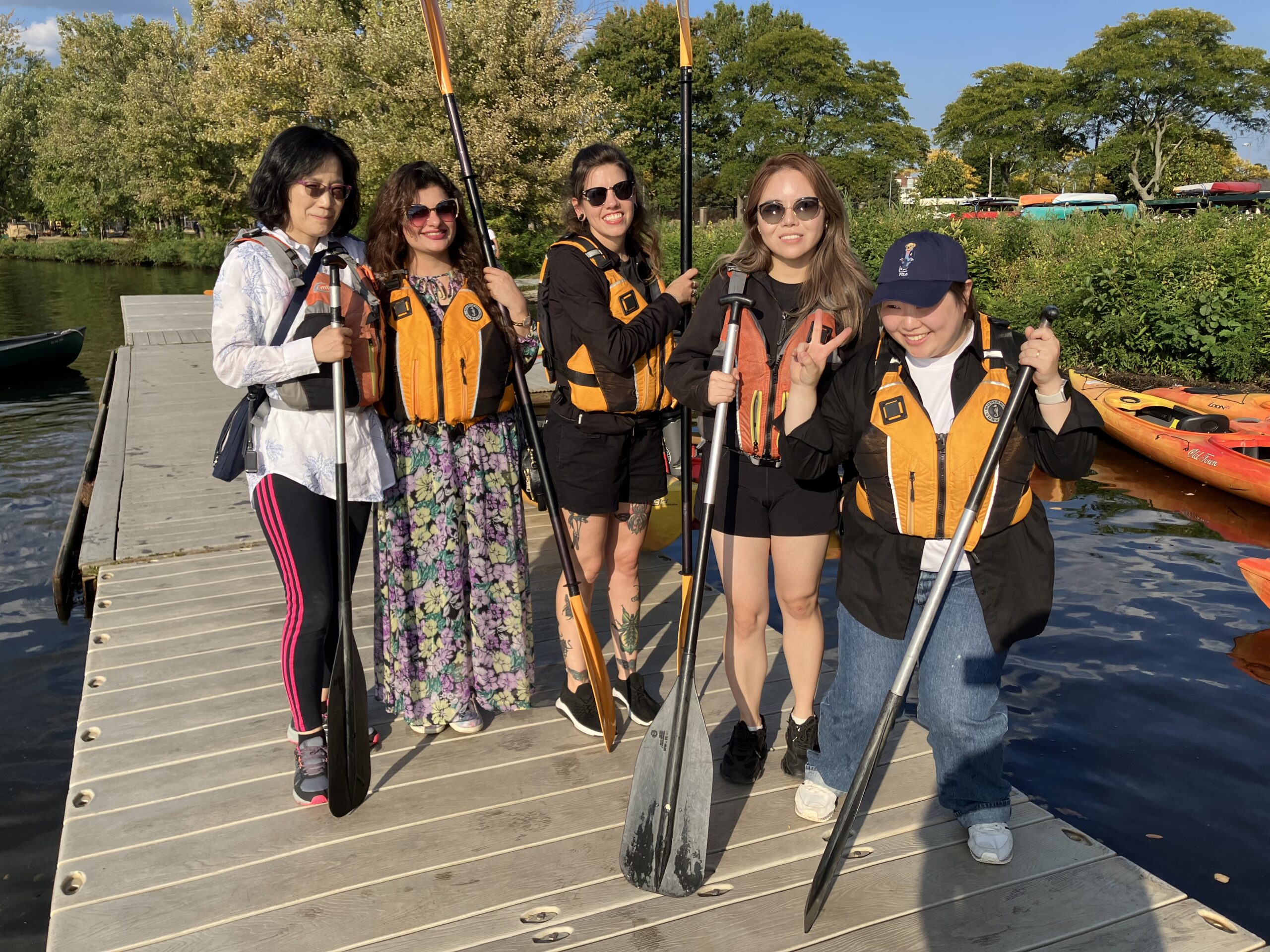 A group of women standing on a river dock with life jackets and paddles.