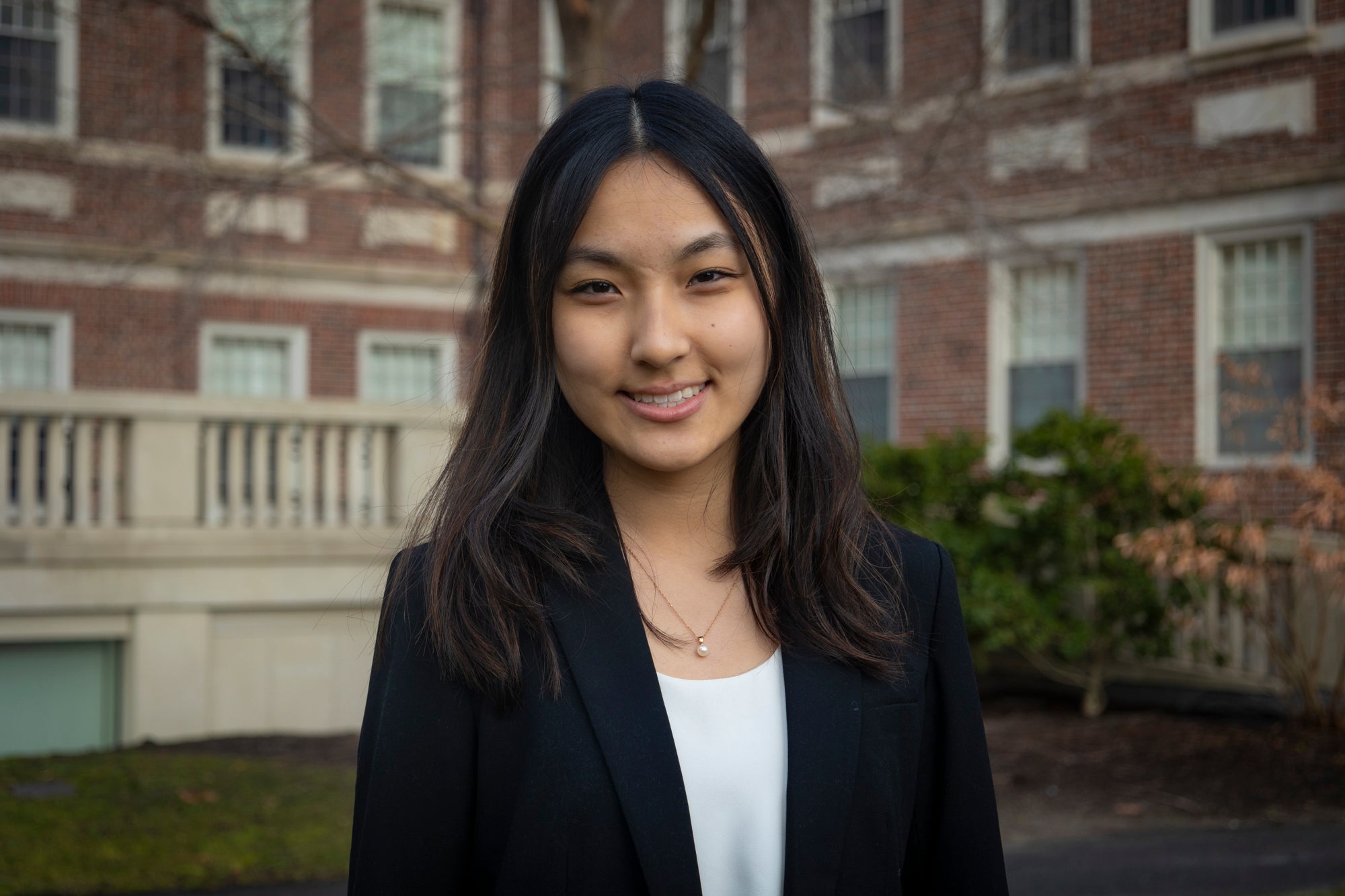 a headshot of Grace Yang. she is outside, and the background is a red brick building with many windows. Grace is smiling at the camera and has her hair down over her shoulders and wears a white blouse under a black blazer. she wears a necklace with a single pearl on the gold chain
